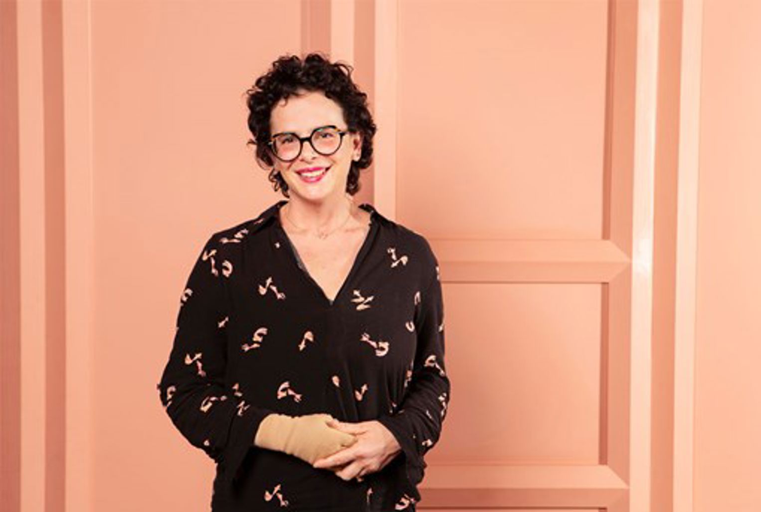 Photo of a smiling woman (Dr Na'ama Carlin) who is wearing glasses and pink lipstick. Her hair is short, dark and curly and she is wearing a dark dress, against a light pink background